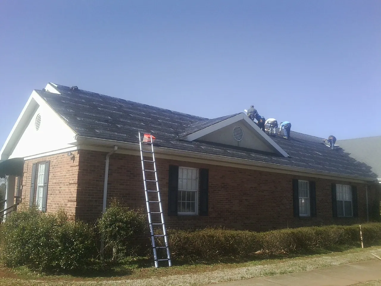 Crew tearing shingles off a brick ranch roof with a ladder set — Metal Roof Repair work by our crew in Simpsonville
