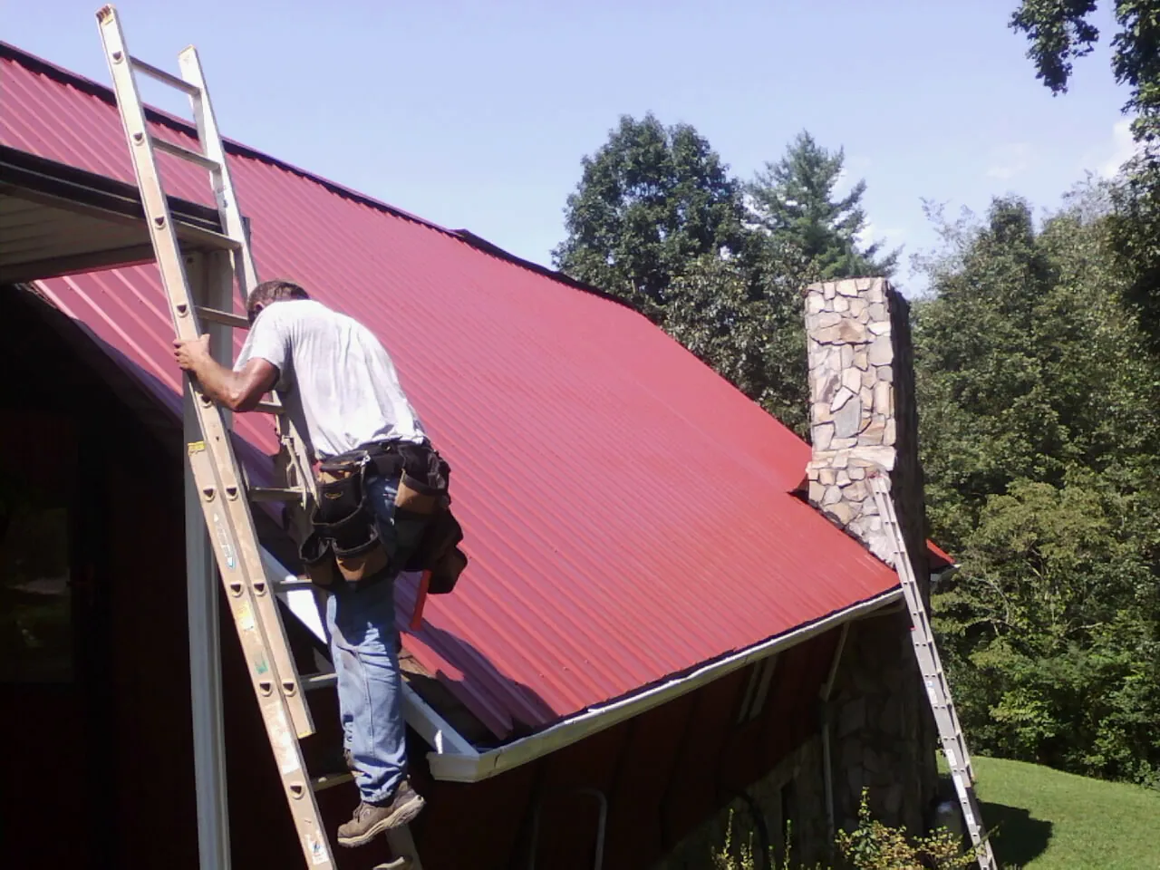 Roofer on a ladder beside a red metal roof and stone chimney — Soffit & Fascia Repair by Roofing Brothers in Simpsonville
